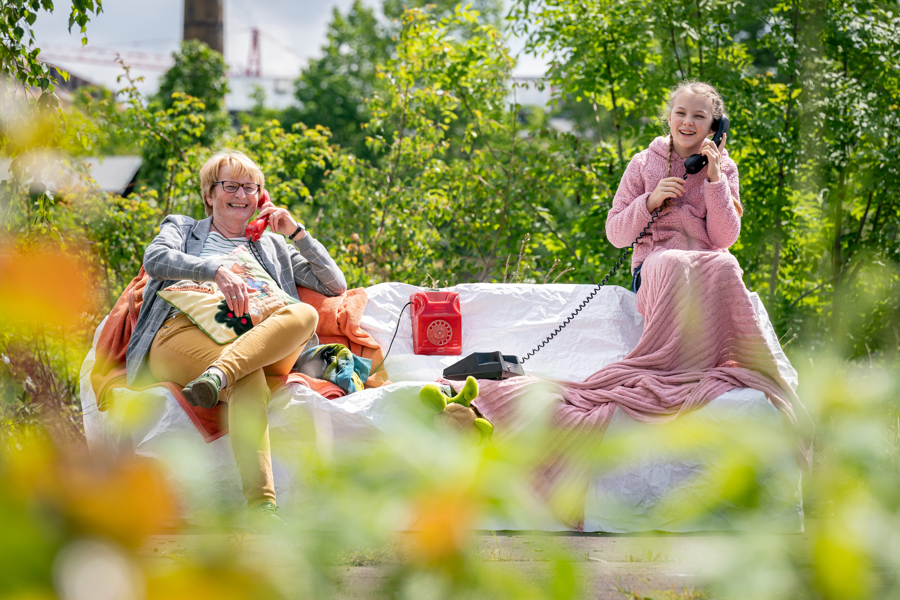 Inszeniertes Foto. Eine ältere Spielerin und eine junge Spielerin sitzen im Freien auf einer Brachfläche mit vielen grünen Pflanzen um sich herum auf einem Sofa. Sie haben maximal viel Abstand voneinander und halten jeweils einen Telefonhörer in der Hand. Während ihrem Gespräch lächeln sie fröhlich in die Kamera. Das Sofa ist aus weißem Papiertuch geformt.